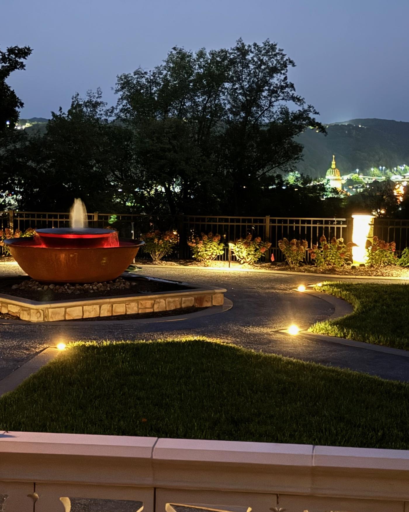 A copper fountain and lit gravel path at dusk, with the valley lights beyond