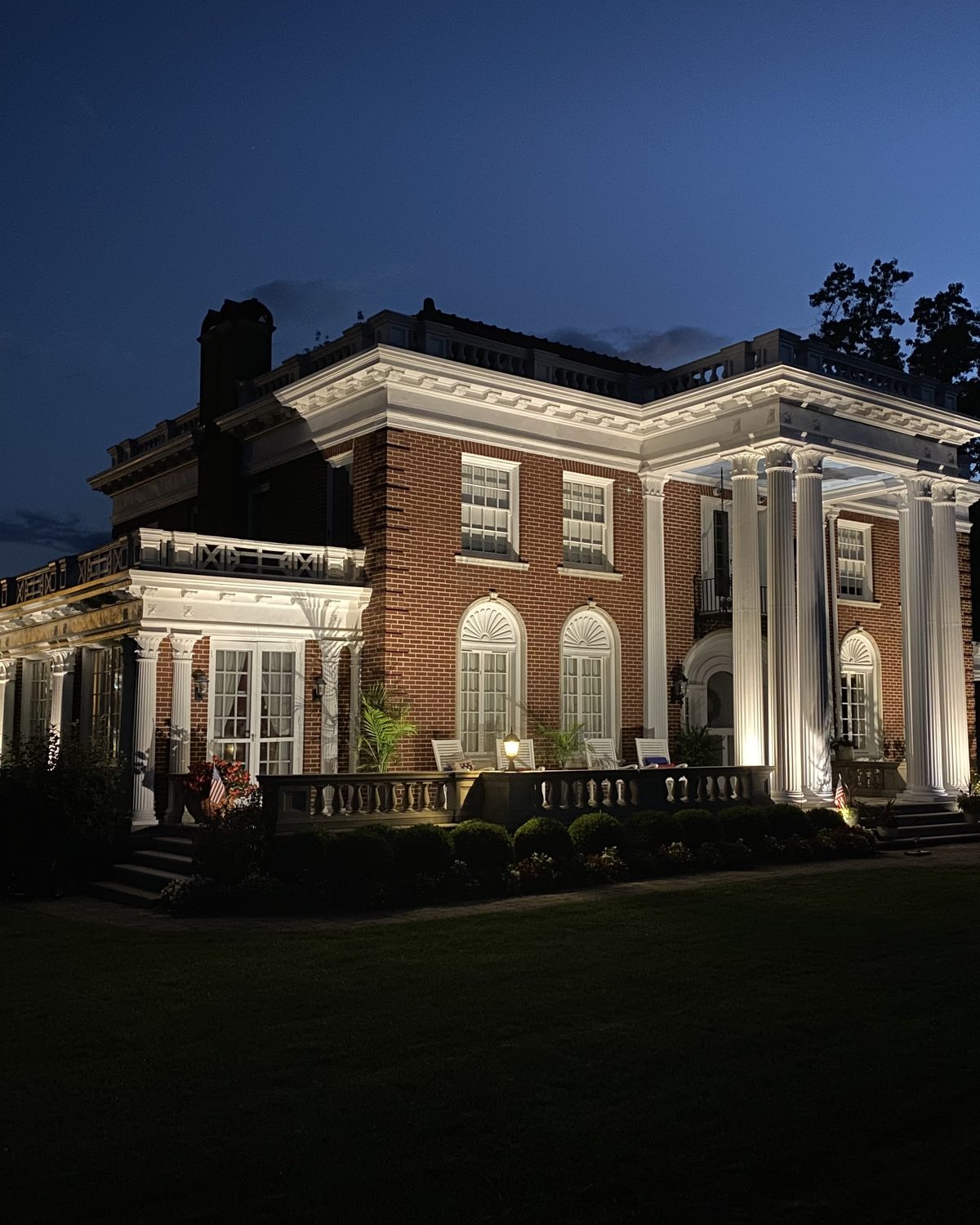 A brick residence lit at dusk, with clipped hedging along the terrace