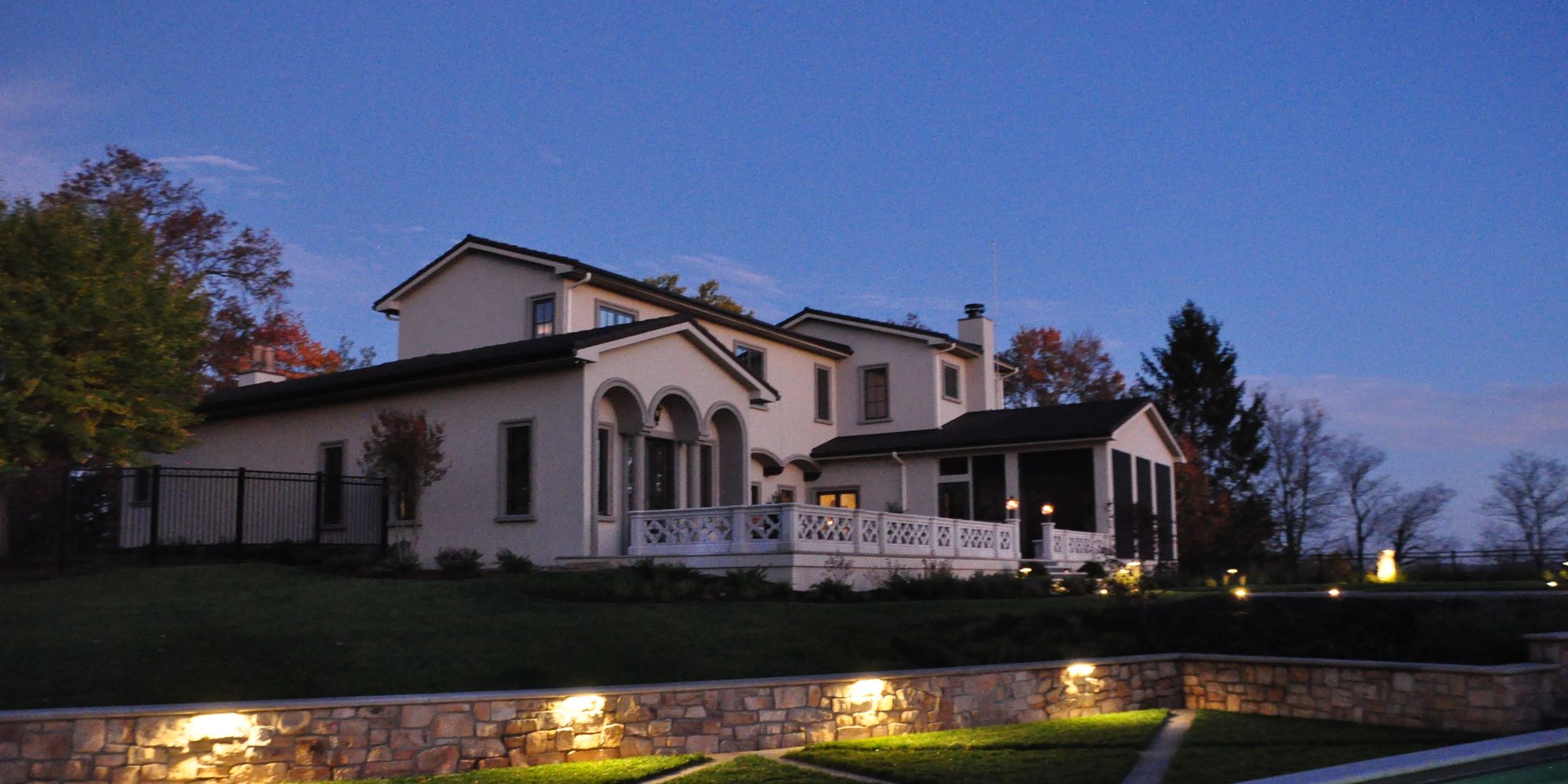 A stucco residence at dusk above a lit stone retaining wall and terraced lawn panels