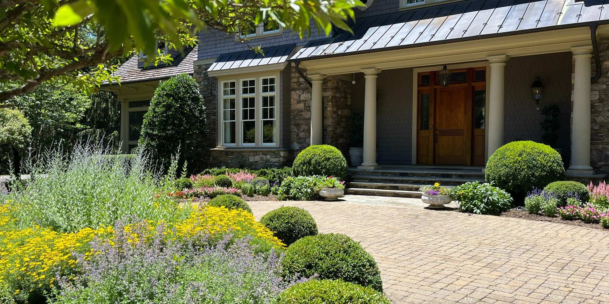 A shingle-and-stone residence with clipped boxwood, layered perennial borders and a paver entrance drive