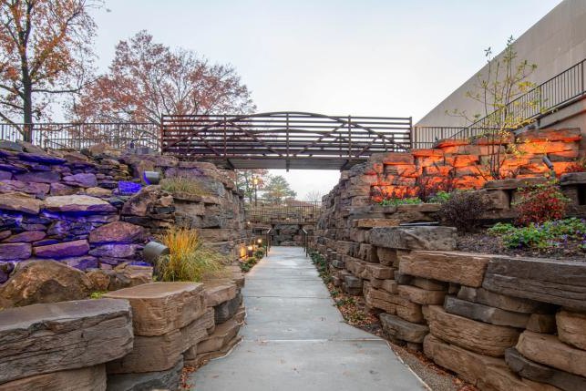 Stacked stone terraces lit in purple and amber, either side of a paved walkway beneath a pedestrian bridge